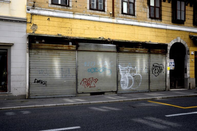 Facciata urbana gialla con saracinesche chiuse, graffiti e ingresso ad arco su strada deserta.