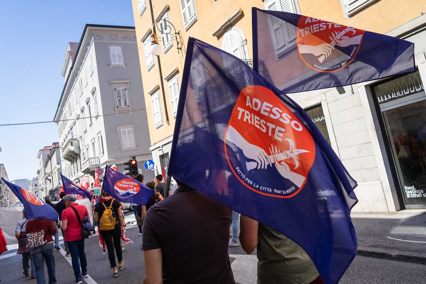 Corteo Adesso Trieste nel centro città con bandiere blu e rosse al sole.