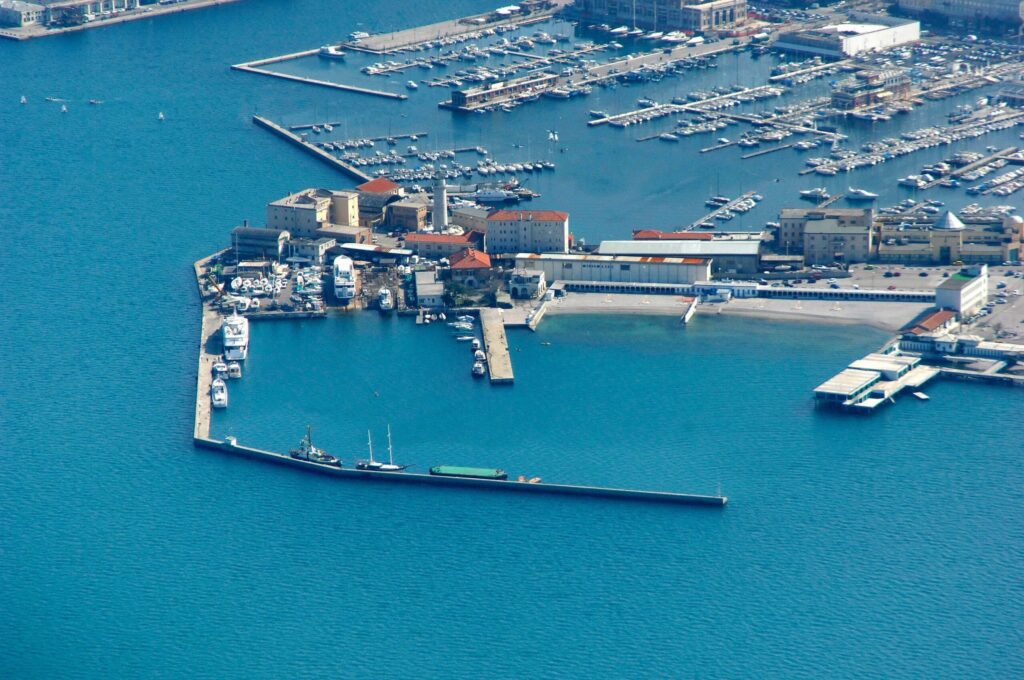 Vista aerea di porto turistico con marina, yacht ormeggiati e frangiflutti sul mare turchese.