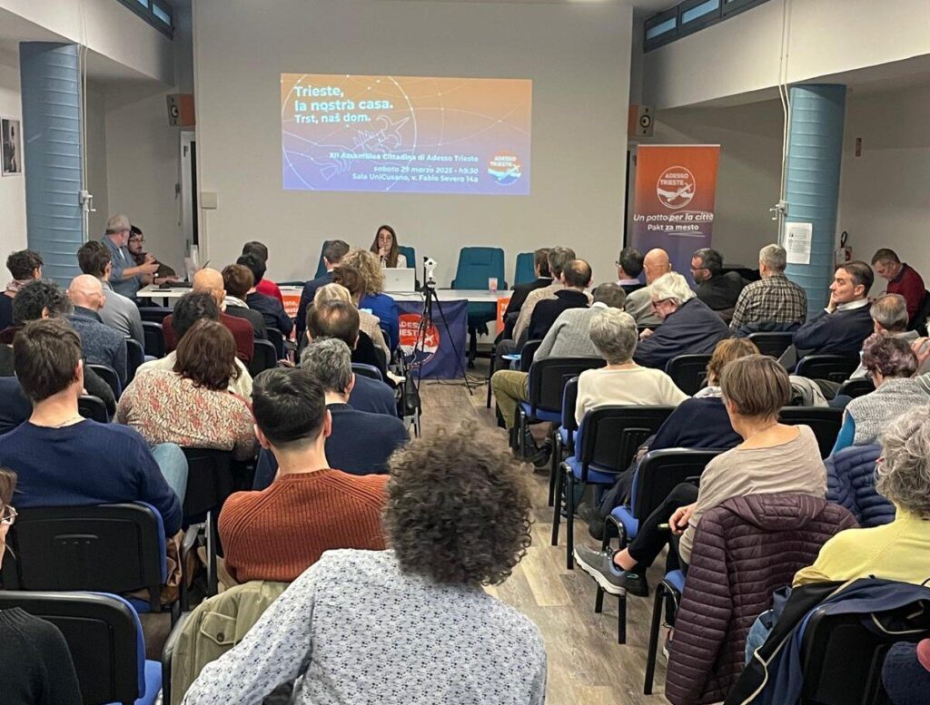 Assemblea cittadina a Trieste con pubblico e relatori durante un dibattito in auditorium