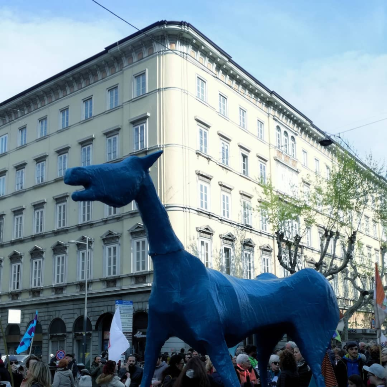 Giraffa blu gigante in piazza storica, tra folla, bandiere e palazzi eleganti.