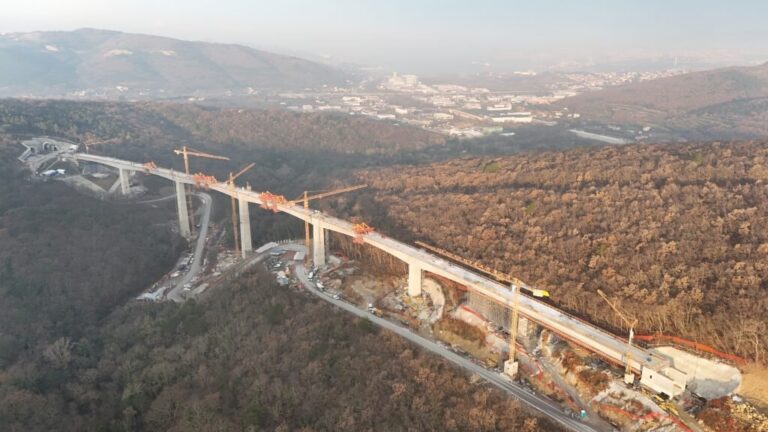 Viadotto in costruzione tra colline boscose, con piloni in cemento e mezzi al lavoro.