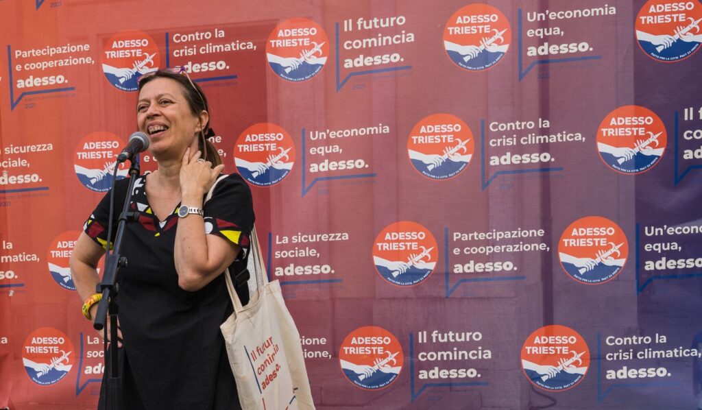 Donna sorridente parla al microfono davanti al backdrop politico “Adesso Trieste” a Trieste.