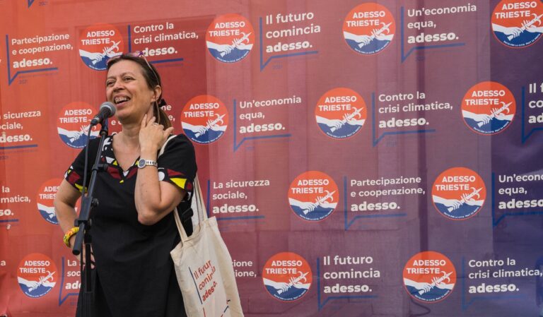 Donna sorridente parla al microfono davanti al backdrop politico “Adesso Trieste” a Trieste.