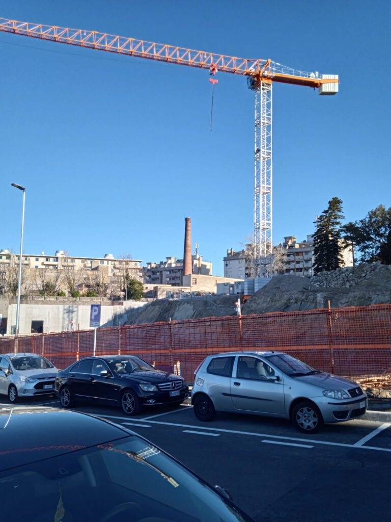 Cantiere urbano con gru, scavi recintati e auto parcheggiate sotto cielo sereno.