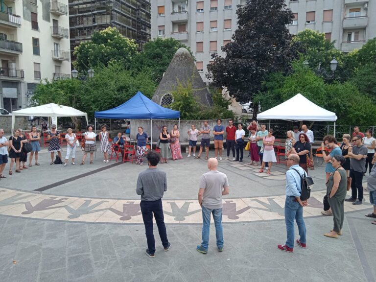Raduno pubblico in piazza urbana con tende informative, persone in cerchio e grattacieli sullo sfondo.