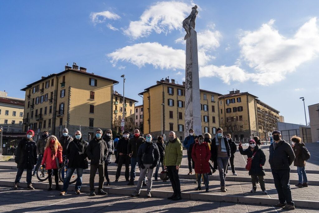 Piazza europea storica con folla, colonna monumentale e palazzi al sole.
