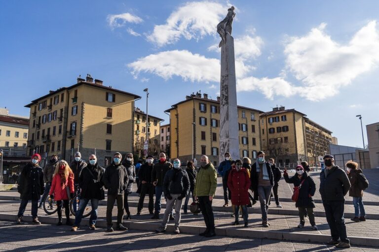 Piazza europea storica con folla, colonna monumentale e palazzi al sole.