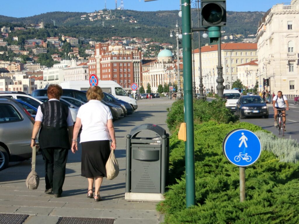 Strada urbana soleggiata con pedoni, auto e panorama collinare storico.