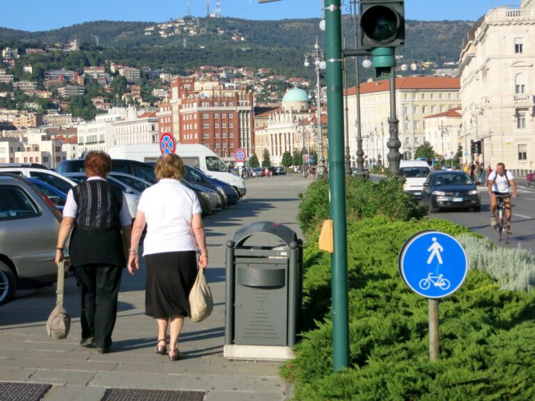 Strada urbana soleggiata con pedoni, auto e panorama collinare storico.