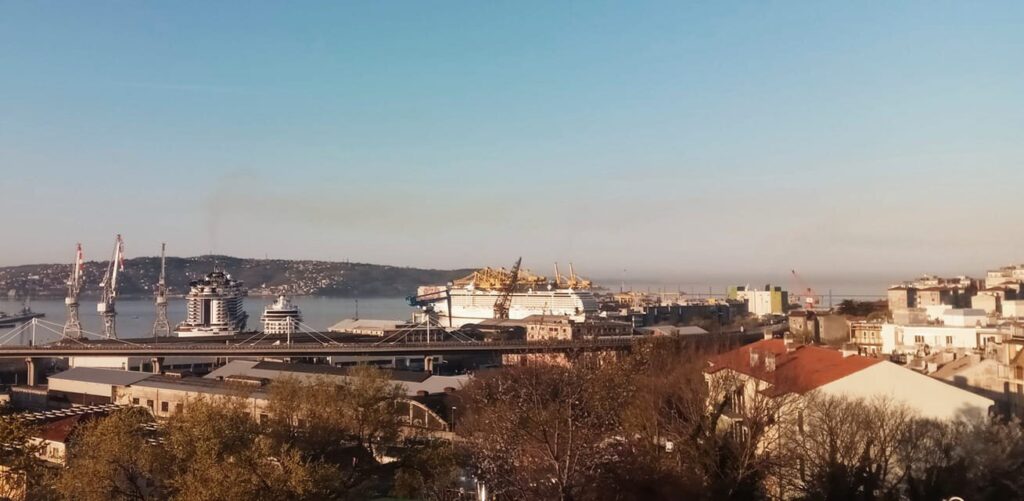 Panorama del porto costiero con gru, traghetto bianco e colline sotto cielo sereno.