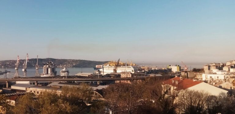 Panorama del porto costiero con gru, traghetto bianco e colline sotto cielo sereno.