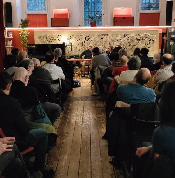 Conferenza intima in sala accogliente con pubblico attento e relatore al tavolo.
