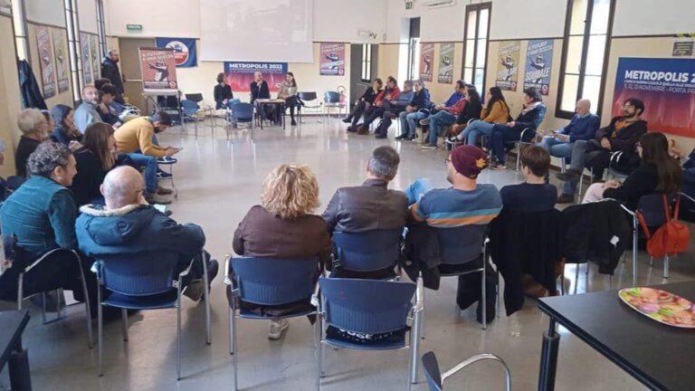 Assemblea di comunità in cerchio, sala luminosa con facilitatore, poster e partecipanti attenti.