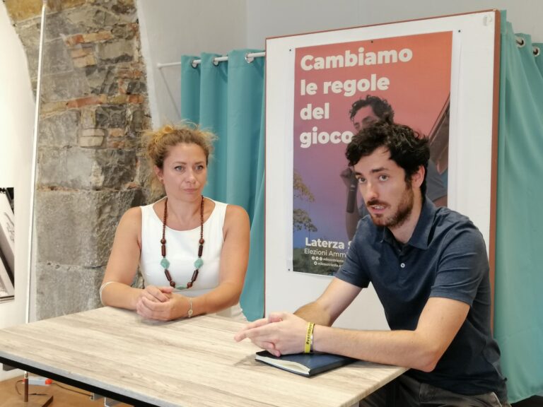 Donna e uomo discutono a un tavolo in sala comunitaria, davanti al poster “Cambiamo le regole del gioco”.