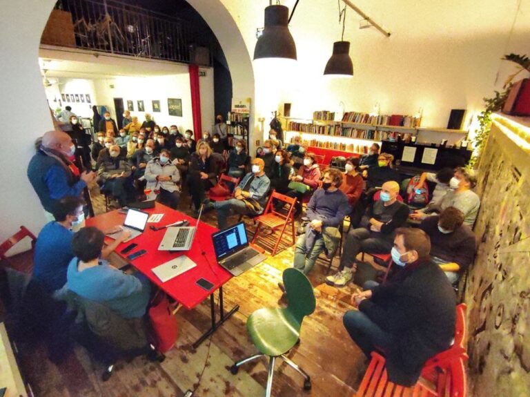 Workshop comunitario in sala rustica con persone attorno a tavoli rossi e laptop.