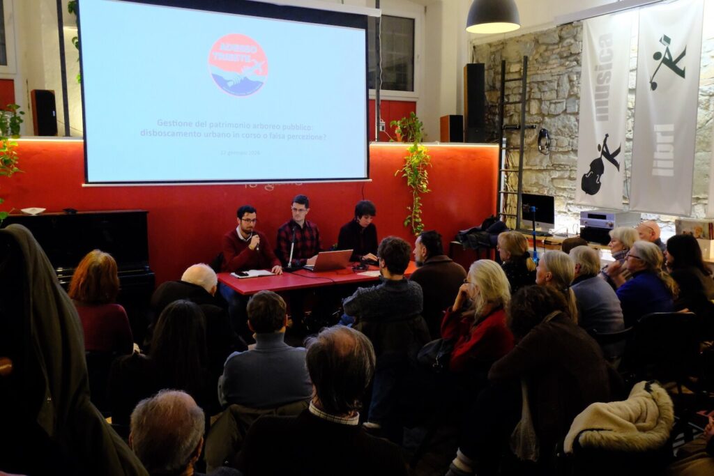 Conferenza pubblica a Trieste con pubblico, relatori al tavolo rosso e proiezione.