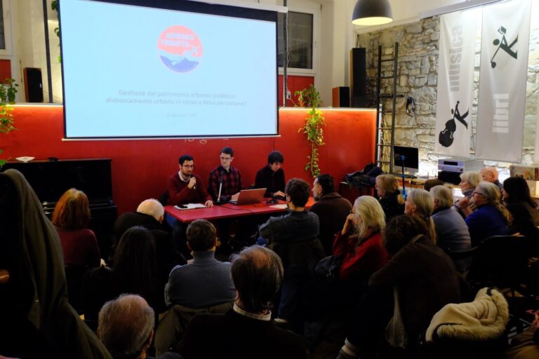 Conferenza pubblica a Trieste con pubblico, relatori al tavolo rosso e proiezione.