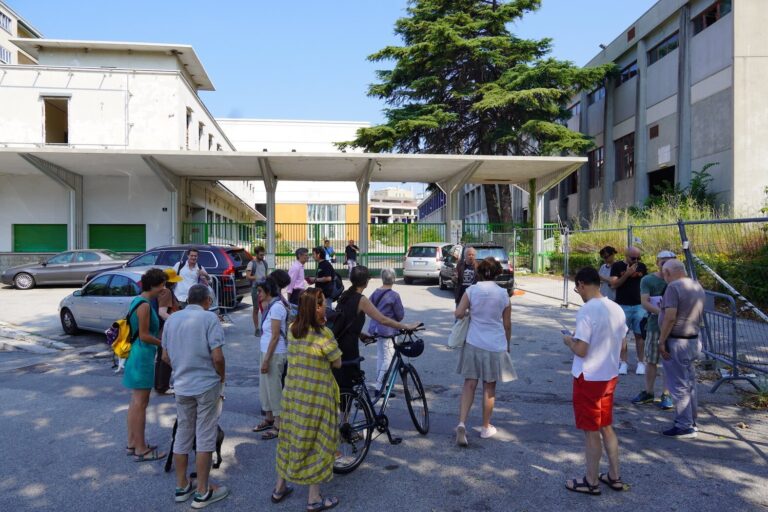Raduno in cortile soleggiato tra edifici con persone, bici e auto parcheggiate.