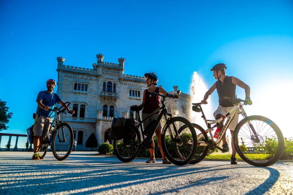Tre ciclisti davanti a un palazzo bianco con fontana e cielo azzurro.