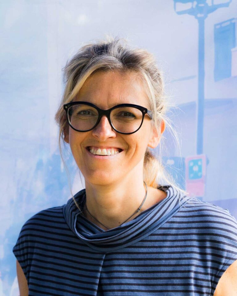 Donna sorridente con occhiali scuri e maglia a righe, ritratto luminoso su sfondo azzurro.