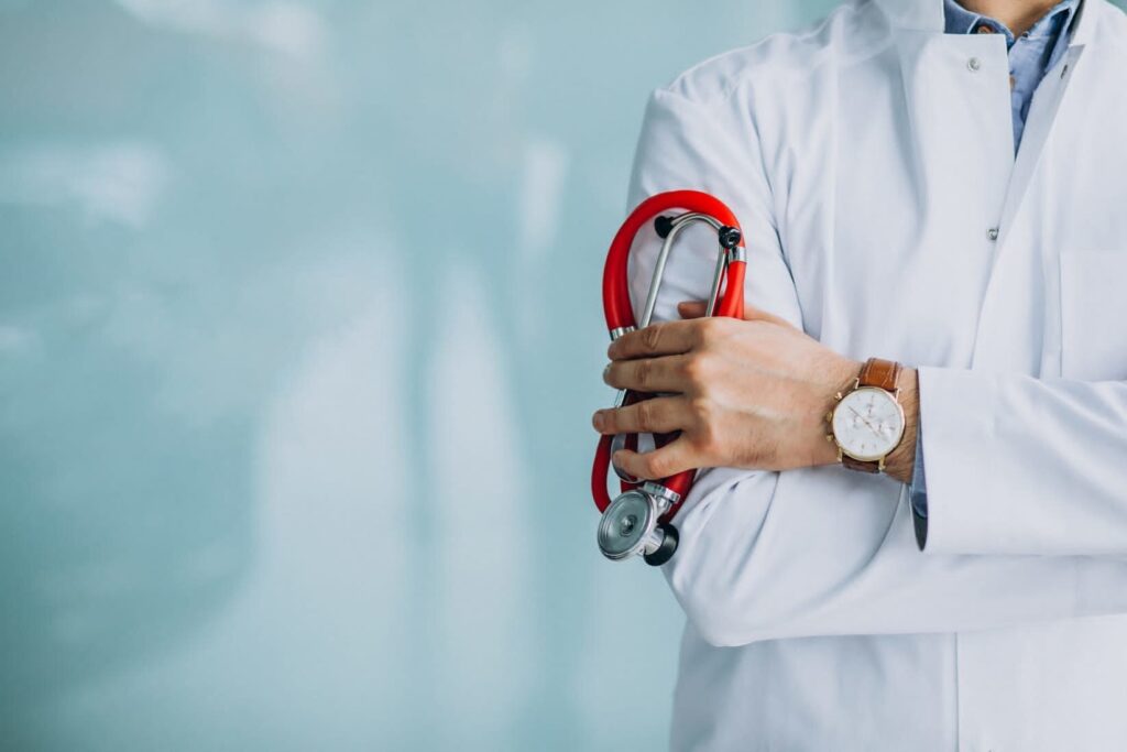 Medico in camice bianco con stetoscopio rosso e braccia incrociate, sfondo clinico sfocato.