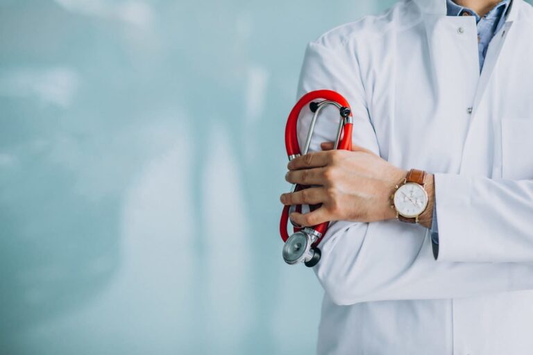 Medico in camice bianco con stetoscopio rosso e braccia incrociate, sfondo clinico sfocato.