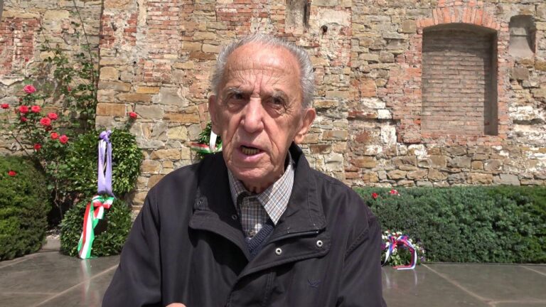 Partigiano anziano alla commemorazione del 25 Aprile, davanti a corone tricolori e muro storico.