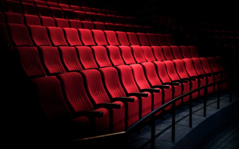 Teatro vuoto con file di poltrone rosse illuminate, atmosfera cinematografica e buia.