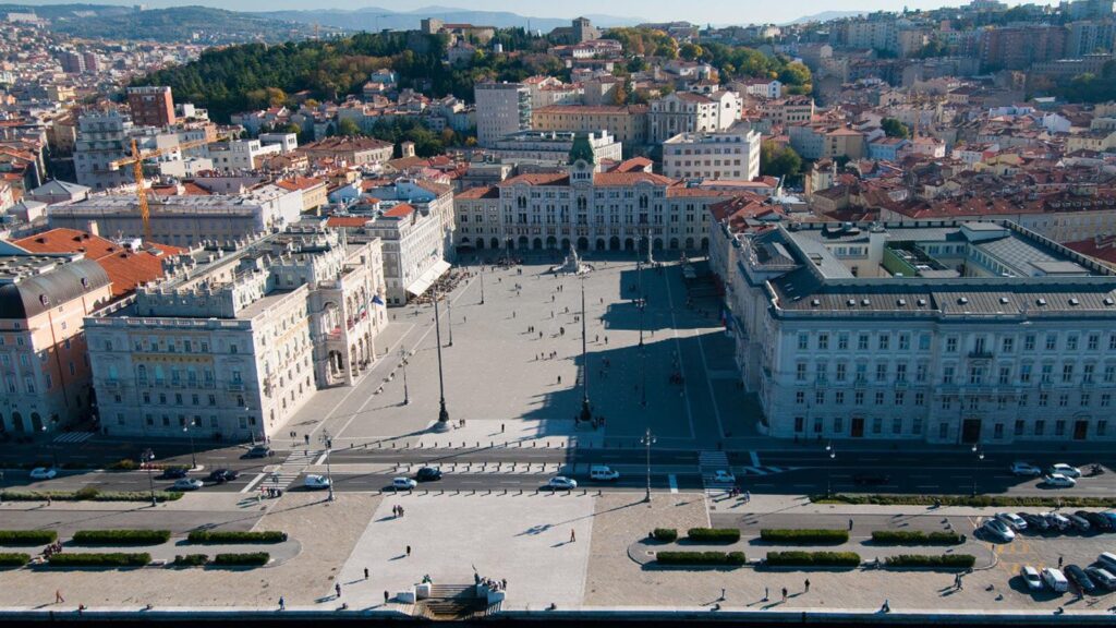 Veduta aerea di piazza europea storica con monumento, palazzi monumentali e città collinare.