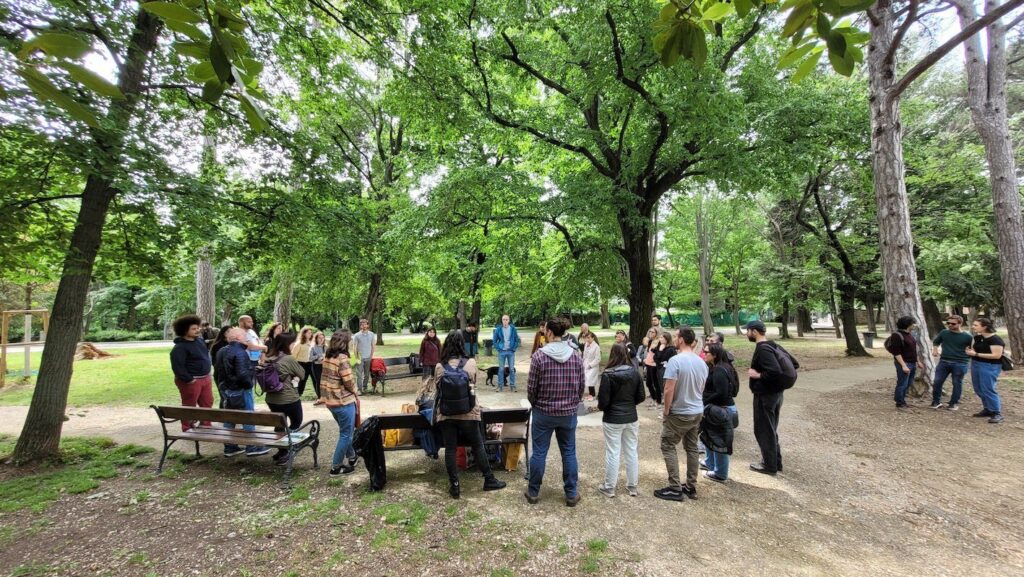 Riunione conviviale nel bosco con tavoli da picnic e persone in radura.