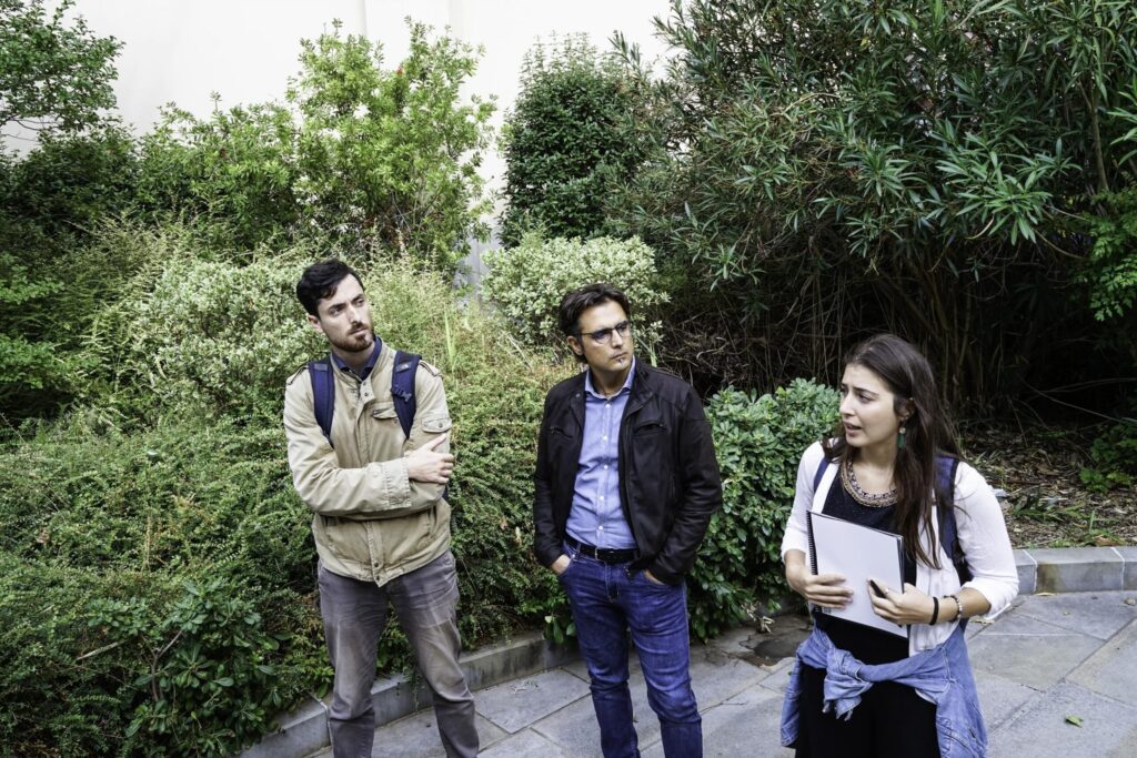 Tre persone con zaini discutono in un giardino verde, mentre una donna mostra appunti.