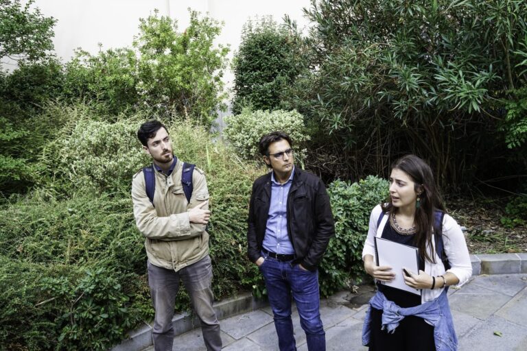 Tre persone con zaini discutono in un giardino verde, mentre una donna mostra appunti.