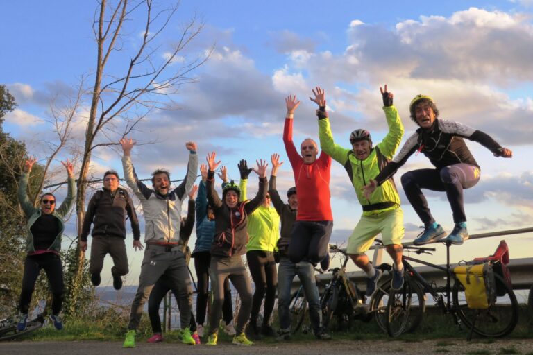 Gruppo di ciclisti festeggia all’aperto con bici, salti e braccia alzate.
