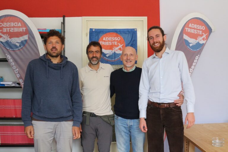 Quattro uomini in ufficio posano davanti a banner promozionali rossi e blu.
