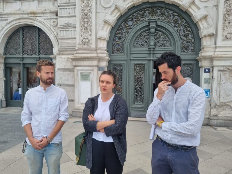 Tre persone discutono davanti a un palazzo storico con archi monumentali.
