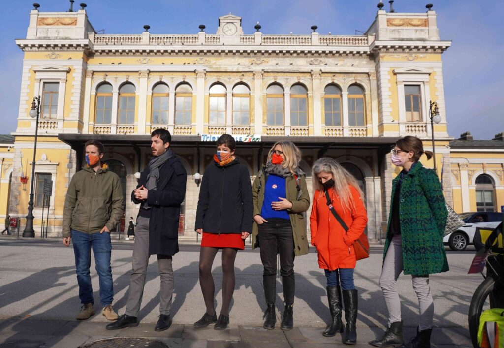 Sei persone in piazza davanti a un palazzo storico, in una giornata soleggiata.