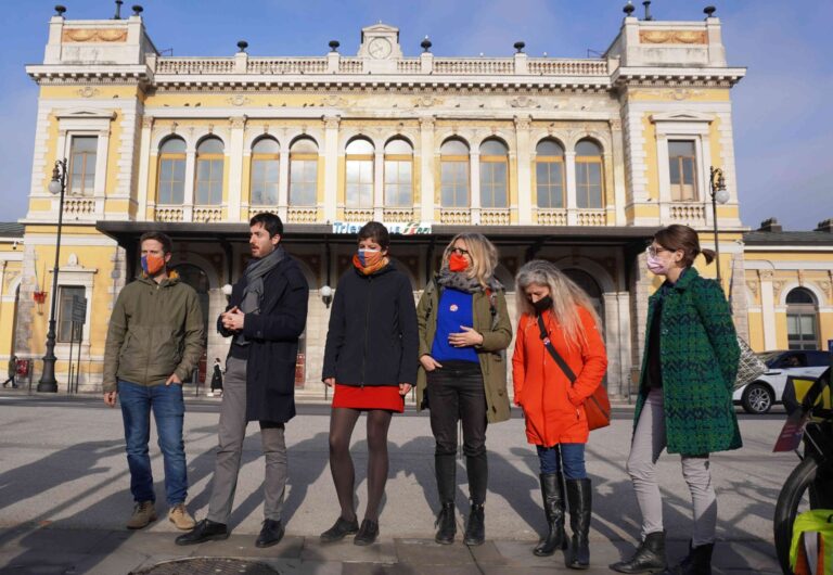 Sei persone in piazza davanti a un palazzo storico, in una giornata soleggiata.