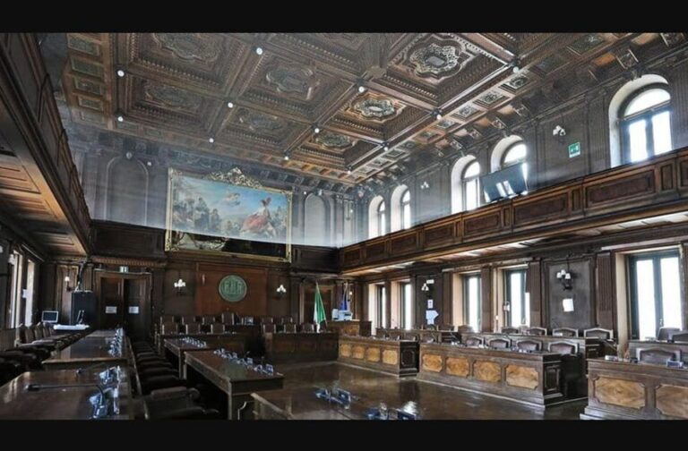 Sala consiliare storica con boiserie, affresco, soffitto cassettonato e banchi in legno.