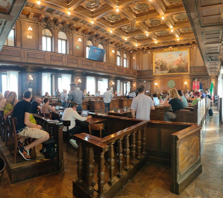 Assemblea pubblica in storica sala consiliare con banchi in legno e bandiere.