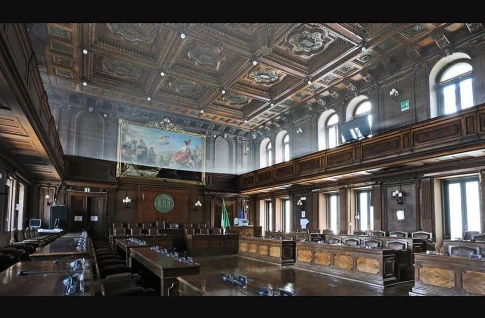 Sala consiliare storica con boiserie, affresco, soffitto cassettonato e banchi in legno.