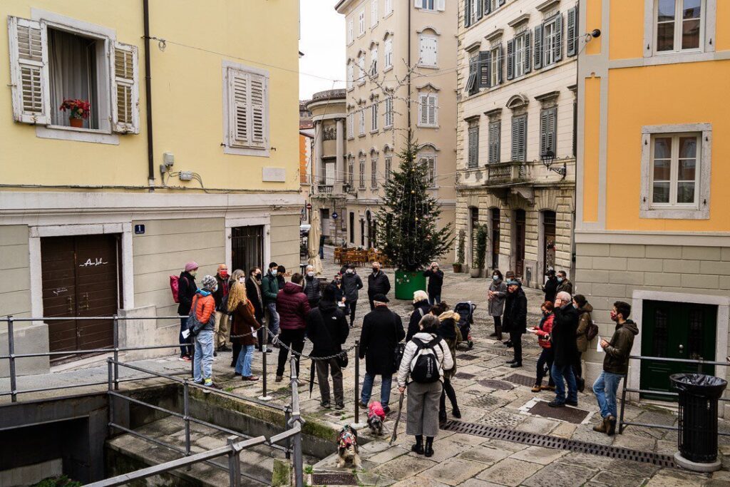 Raduno di persone in una stretta via storica europea con edifici pastello e pavé.