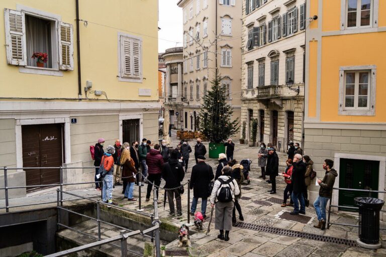 Raduno di persone in una stretta via storica europea con edifici pastello e pavé.