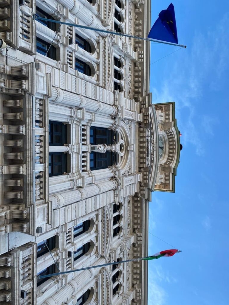 Facciata storica istituzionale in pietra chiara con bandiere, vista dal basso sotto cielo azzurro.