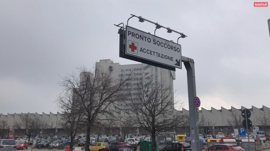 Cartello Pronto Soccorso con freccia verso l’ospedale, auto in città e cielo grigio.