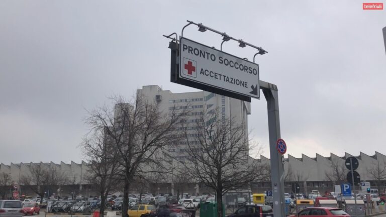 Cartello Pronto Soccorso con freccia verso l’ospedale, auto in città e cielo grigio.