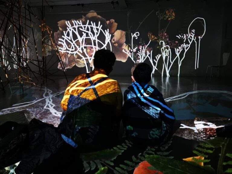 Due spettatori osservano un albero luminoso in un’installazione artistica immersiva.