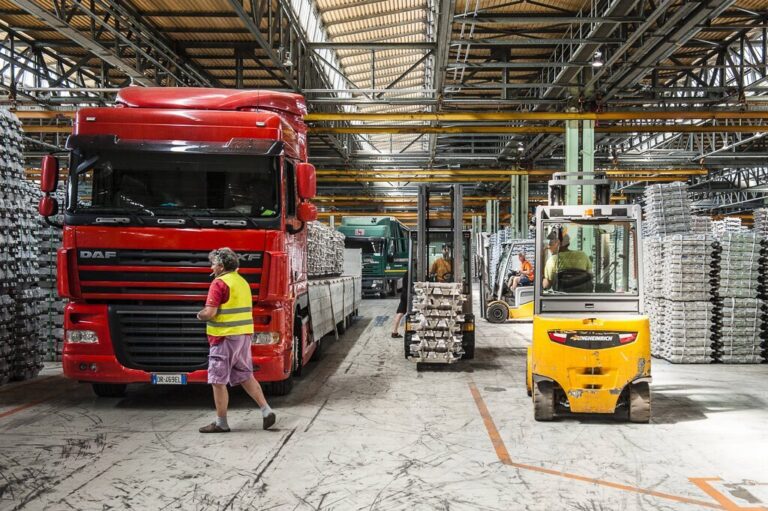 Magazzino industriale con camion rosso, carrello elevatore giallo e operatore in sicurezza.