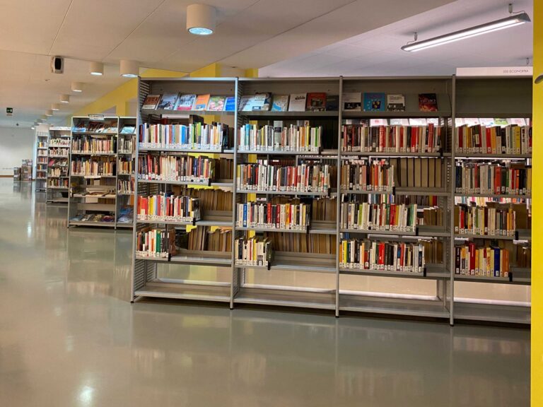 Interno di biblioteca moderna con scaffali metallici pieni di libri e corridoio spazioso.