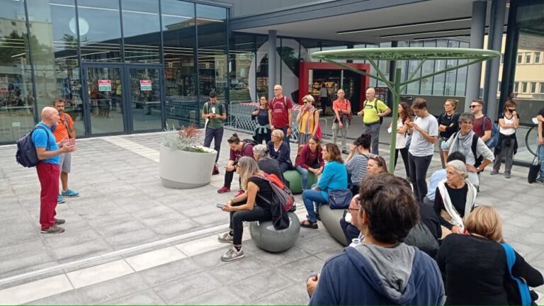 Gruppo riunito davanti a edificio moderno in vetro, guida parla in una piazza urbana.